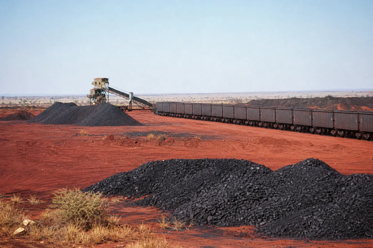Black manganese ore stockpiles on deep red Kalahari sand beside a rail loadout with a line of open wagons.